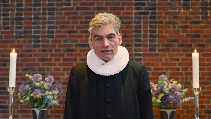 Mand i sort kaptajnjacke og hvid krage står for murvæg med blomster og lygter