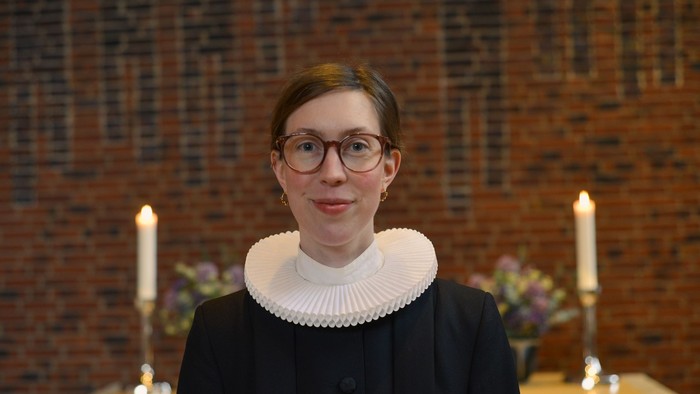 Kvinde i sort og hvid tøj med briller og hårbånd står forud for murvæg med lysstake.