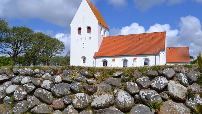 En hvid kirke med rødt tag og tårn bag en stenmur.