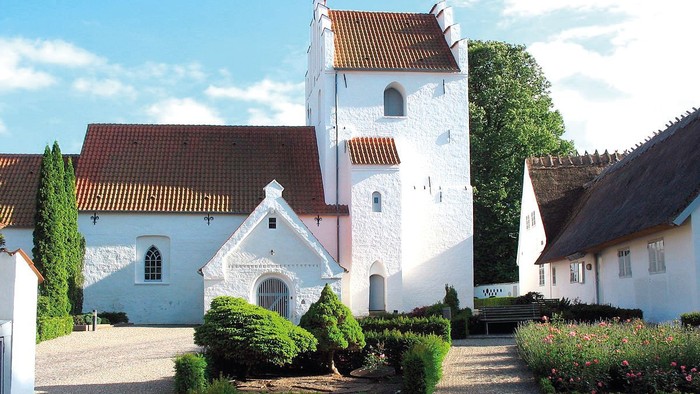 En smuk og hvid kirke med tårn og røde tag i en trædit og grønne omgivelser
