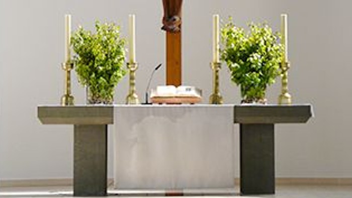 Ein hölzernes Kruzifix steht auf einem Altar mit zwei Kerzenhaltern und Topfpflanzen in einer Kirche.