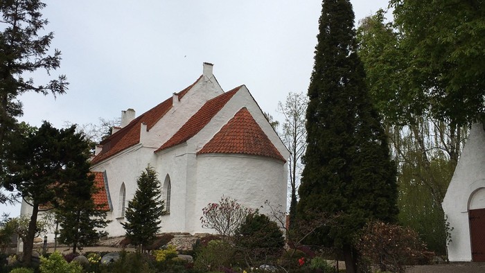 Hvid kirke med rødt tag og kirkegårdens træer i forgrunden