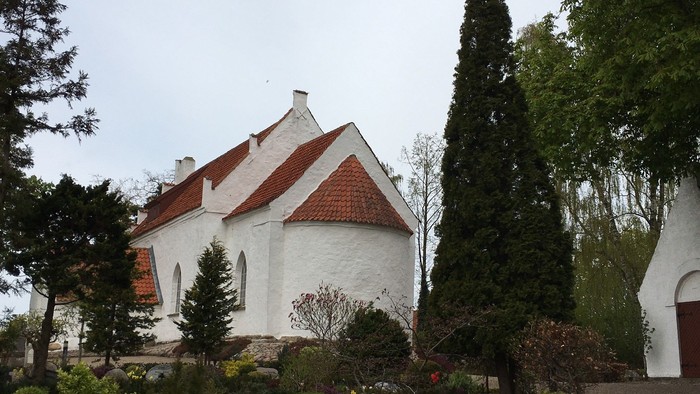 Hvid kirke med rødt tag og kirkegårdens træer i forgrunden