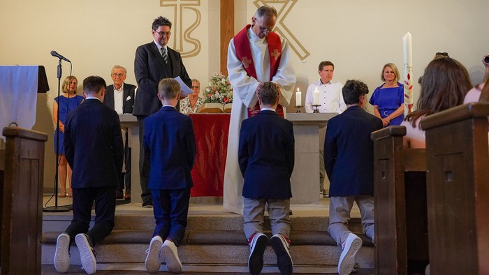 Kinder in Anzügen erhalten Segnungen während einer kirchlichen Zeremonie, geleitet von Geistlichen in traditionellen Gewändern.