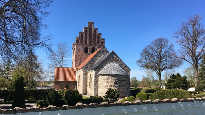 En gammel kirke med tårn og rundtårn i en park med træer og en fontæne.