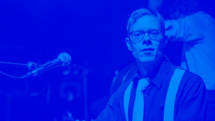 En mand med briller og stribedet skjorte sidder ved et bord med mikrofoner.