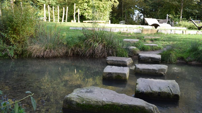 Es ist ein Spielplatz im Grünen zu sehen mit Wasser im Vordergrund. Im Wasser liegen große Steine, die herausragen.