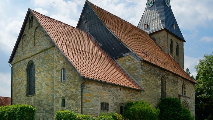 Auf dem Bild sieht man die Evangelische Kirche in Bad Sassendorf Lohne. Sankt Pantaleon. Im Vordergrund stehen grüne Büsche und im Hintergrund sieht man einen hellblauen Himmel mit Schleierwolken. Die Sonne scheint.