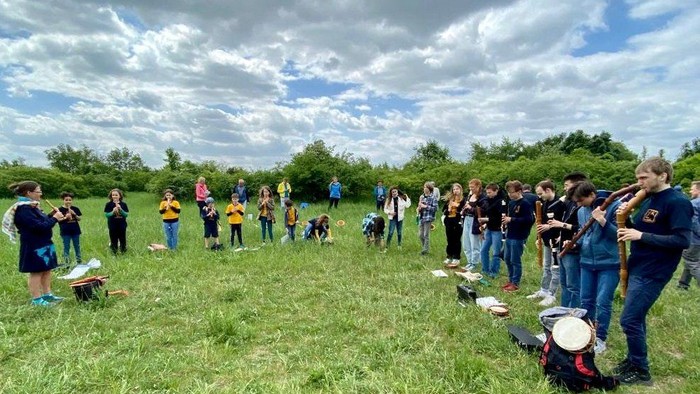 Gruppe von Menschen im Freien, die auf einer grasbewachsenen Wiese unter einem bewölkten Himmel verschiedene Musikinstrumente spielen.