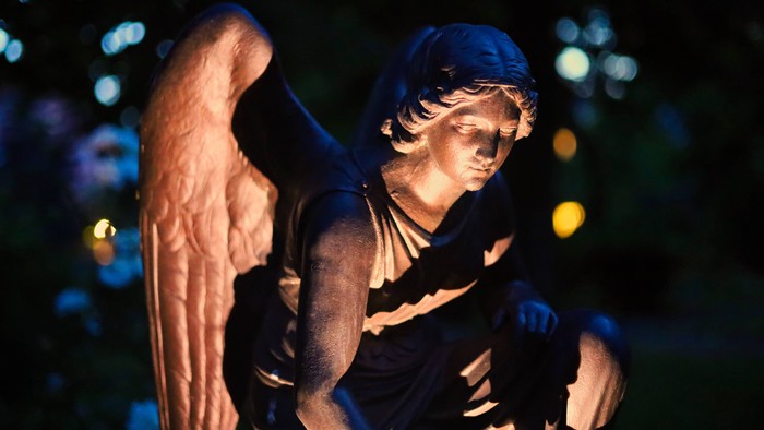 Eine feierliche Engelsstatue mit ausgebreiteten Flügeln in einer schwach beleuchteten, nächtlichen Umgebung.