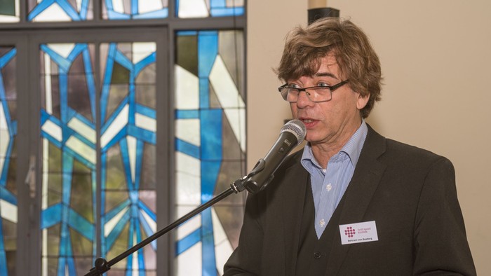Ein Mann im Anzug spricht in ein Mikrofon bei einer Indoor-Veranstaltung mit farbigem Kirchenfenster im Hintergrund.
