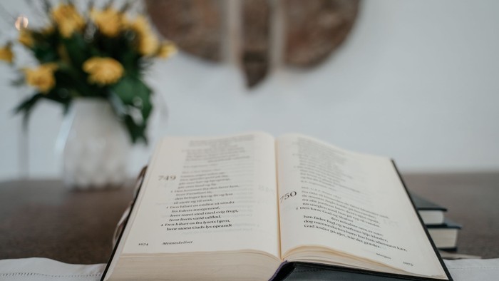 En åben bog på bord med blomster i vaase og klokke på væg i baggrunden