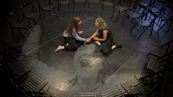 To kvinder sidder i en kreds af stoler og holder hånd.