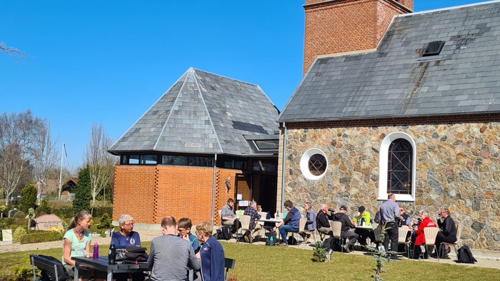 En gruppe af mennesker sidder udendørs ved en kirke og spiser.