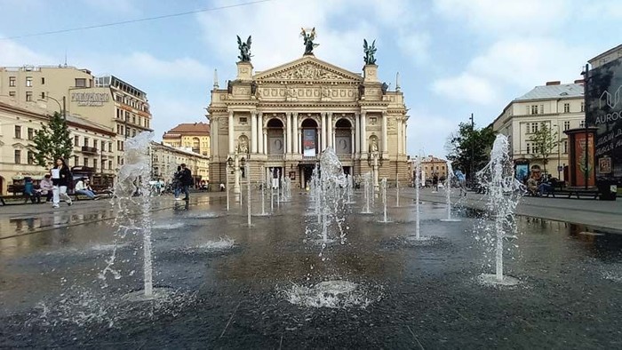 Das Bild zeigt einen Brunnen vor einem großen, prächtigen Gebäude mit Statuen und Springbrunnen im Vordergrund.