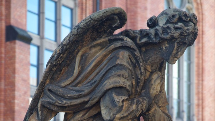 Die Bronzestatue eines geflügelten Wesens vor einem Backsteingebäude.