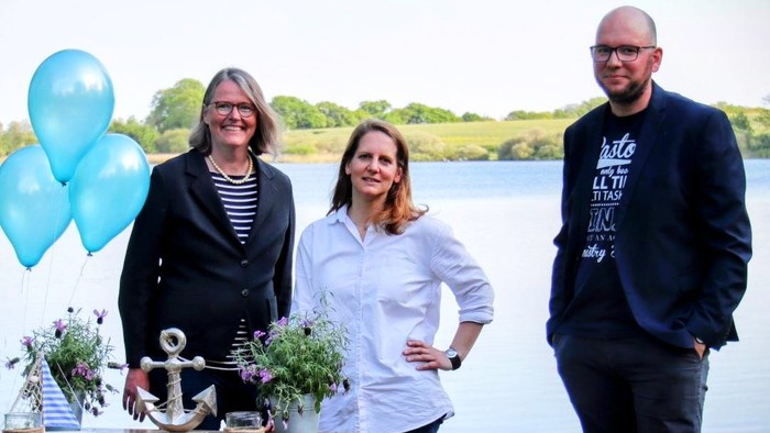 Drei Personen stehen an einem Tisch mit Brot und Ballons am See