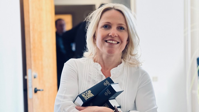 En kvinde med blond hår holder en mørk bog og smiler.