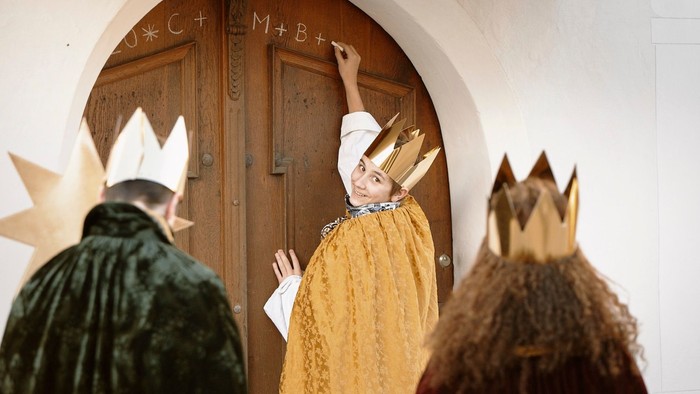 Ein Kind in goldener Krone klopft an eine Holztür, während zwei andere Kinder in Königskostümen danebenstehen.