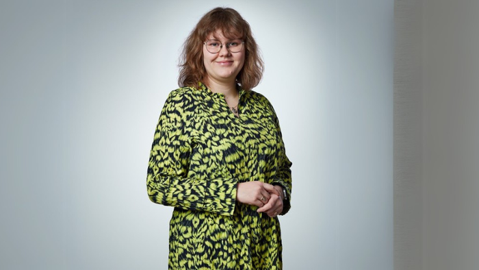 En kvinde står mod en gul væg i en grøn og sort fløjtefarvet jakke.