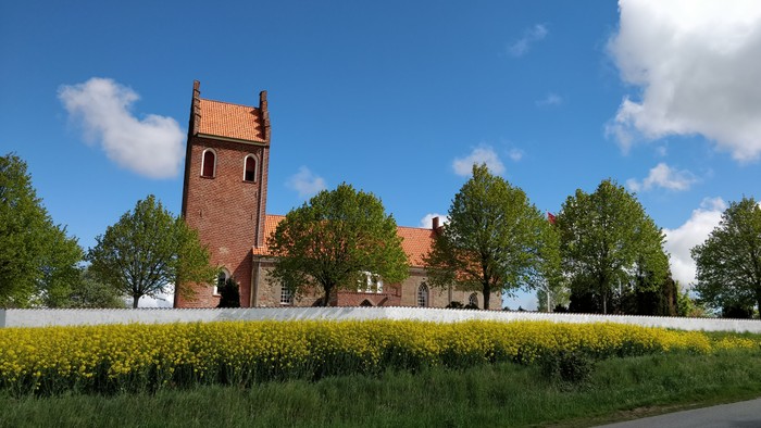 En rød kirke med tårn og klokke, omgivet af grønne træer og gule blomster.