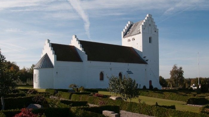 Et hvidt kirkebygningskompleks med tårn og skærmvandret på grønne arealer