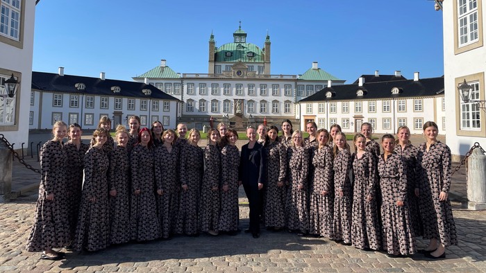 En gruppe kvinder i mønstrede kjoler står sammen foran et historisk slot.