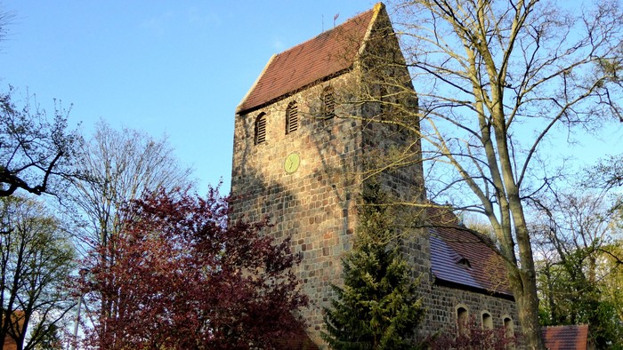 Alte Kirche mit hohem Turm und rotem Dach, umgeben von Bäumen und Büschen