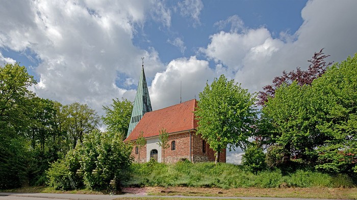 Kleine Kirche mit hohem Turm, umgeben von Bäumen und Büschen, an einer Straße
