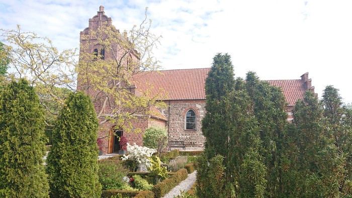 Et gammelt teglsteinshus med rødt tag og en have med træer og buske.
