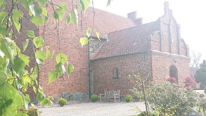 En gammel kirke med teglmur og spisse tårn, omgivet af græs og blomster.