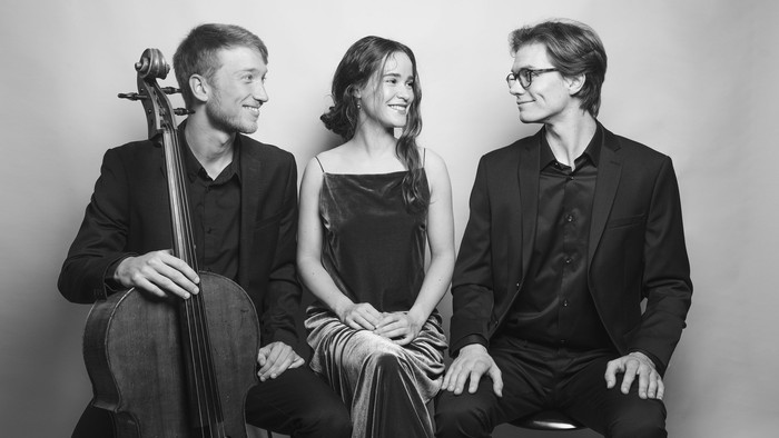 En trio af musikere poserer sammen, en spiller cello, en sitter ved et piano og en står ved siden af dem.