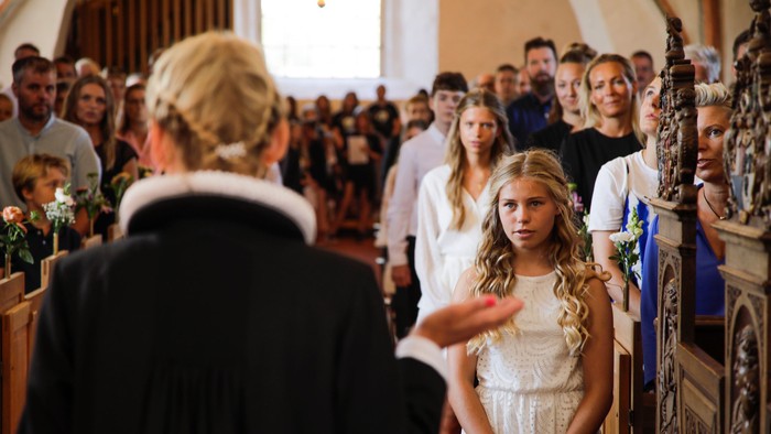 En pige i hvidt tøj deltager i en religiøs ceremoni i en kirke, mens en præst taler til hende.