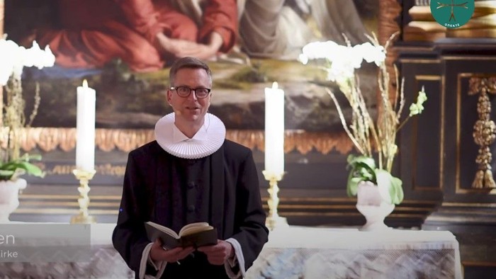 Mand i svart kapittel i kirke med bok og blomsterstykke.