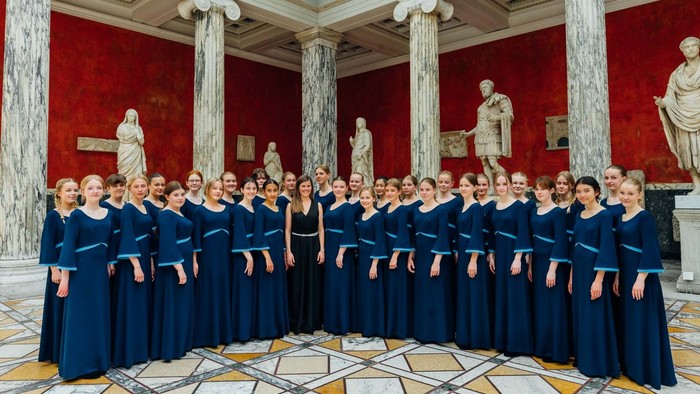 En gruppe kvinder i blå koredragter står sammen i et rom med marmorkolonner og oldtidsskulpturer.
