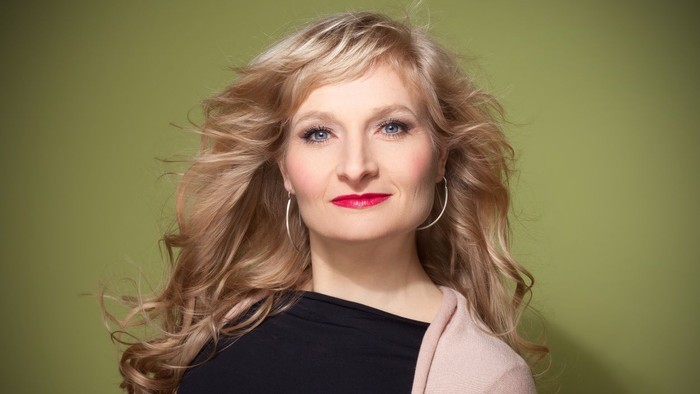 Eine Frau mit langen blonden Haaren, die ein schwarzes Oberteil und eine hellrosa Strickjacke trägt, posiert selbstbewusst.