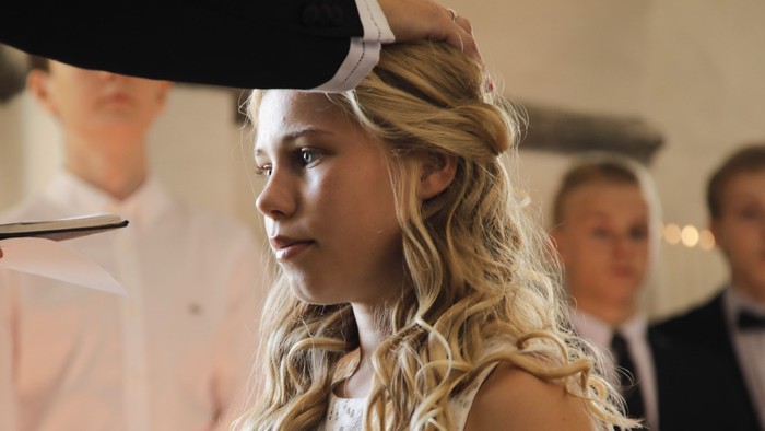 En kvinde med blond hår får sit hår stillet af en frisør, mens to mænd i formel klædning ser til.