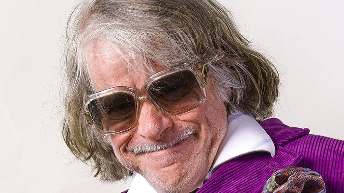 Ein älterer Mann mit langen grauen Haaren und Sonnenbrille lächelt.