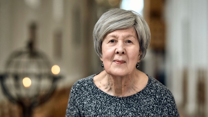 En eldre kvinde med kort grå hår, ståer i et rum med blå vinduer og belysning.