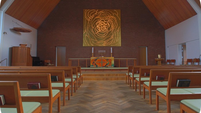 Indenrigsrom med træbænke og alter med stor guldfarvet maleri.