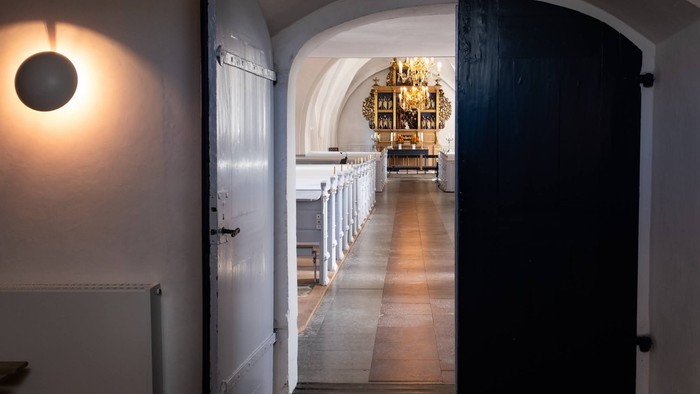 En lang, smal korridor med bænke til venstre og dør til højre, med lysende kælderlys og gulv med mønstre.