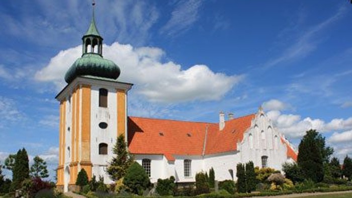 Et hvidt kirkebygning med orange tag og en tårn med grøn kuppel, omgivet af græs og træer under en blå himmel