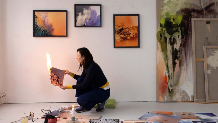 Künstlerin arbeitet in ihrem Atelier an einem Gemälde, umgeben von Kunstwerken und Materialien.