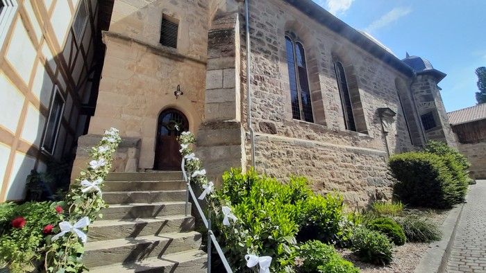 Steingebäude mit Rundbogenfenstern und -tür, umgeben von Grünflächen und Blumen.