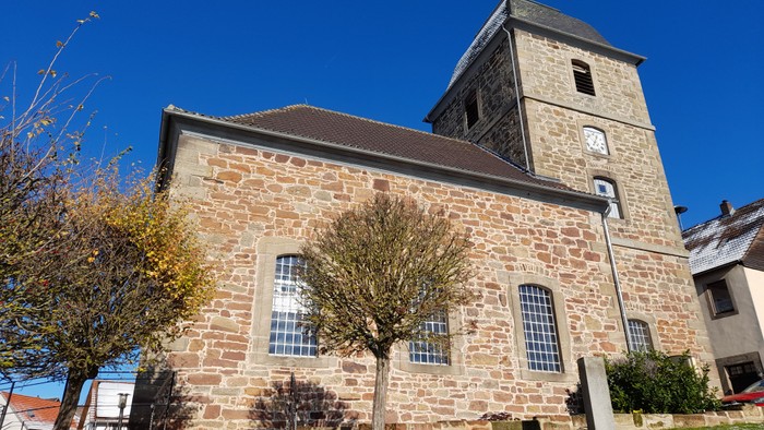 Steinerne Kirche mit hohem Turm und Uhr, umgeben von Bäumen und klarem blauem Himmel.