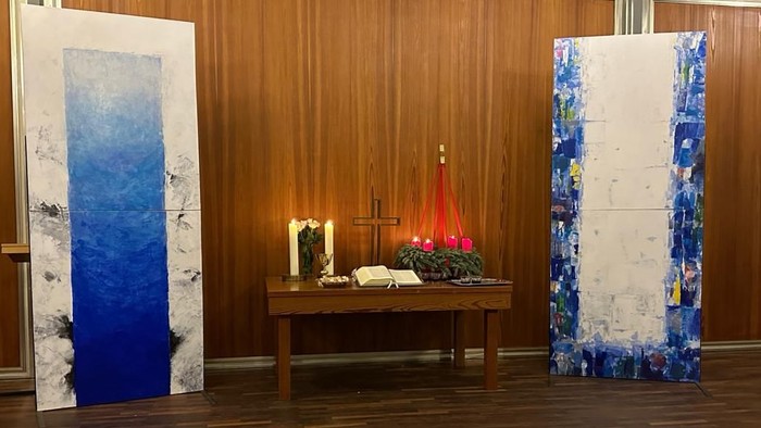 Zwei abstrakte Gemälde flankieren einen kleinen Altar mit einer roten Kerze und einem Kreuz.