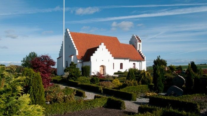 En hvid kirke med rødt tag og en flagstang foran, omgivet af grønne planter og buske