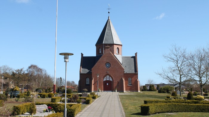 En rød kirke med spids tårn og klokke, beliggende i en park med sti og belysning.