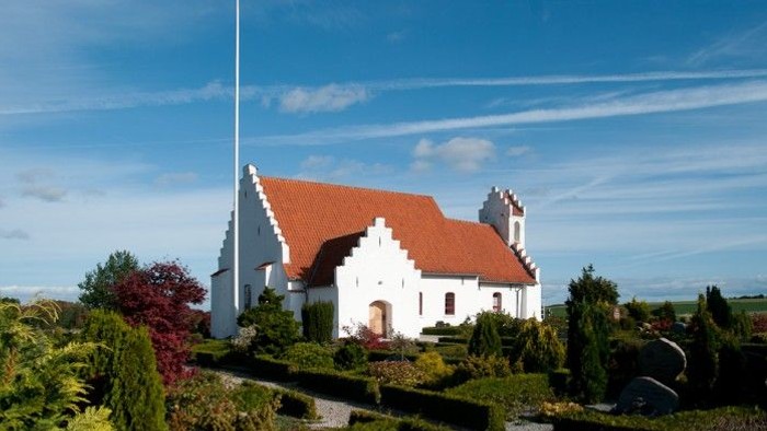 En hvid kirke med rødt tag og en flagstang foran, omgivet af grønne planter og buske