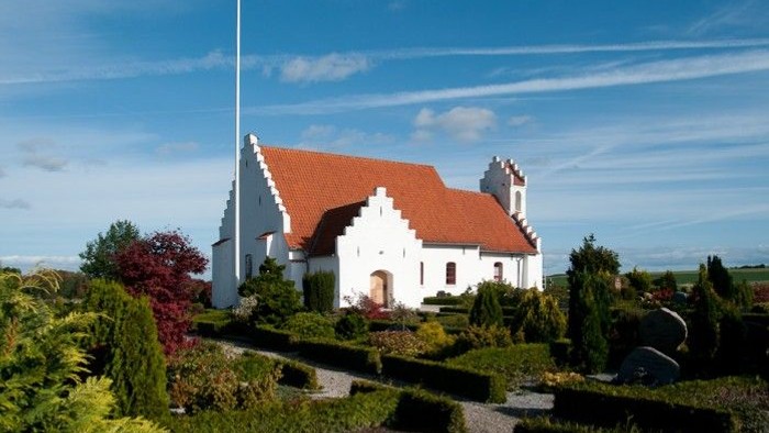 En hvid kirke med rødt tag og en flagstang foran, omgivet af grønne planter og buske